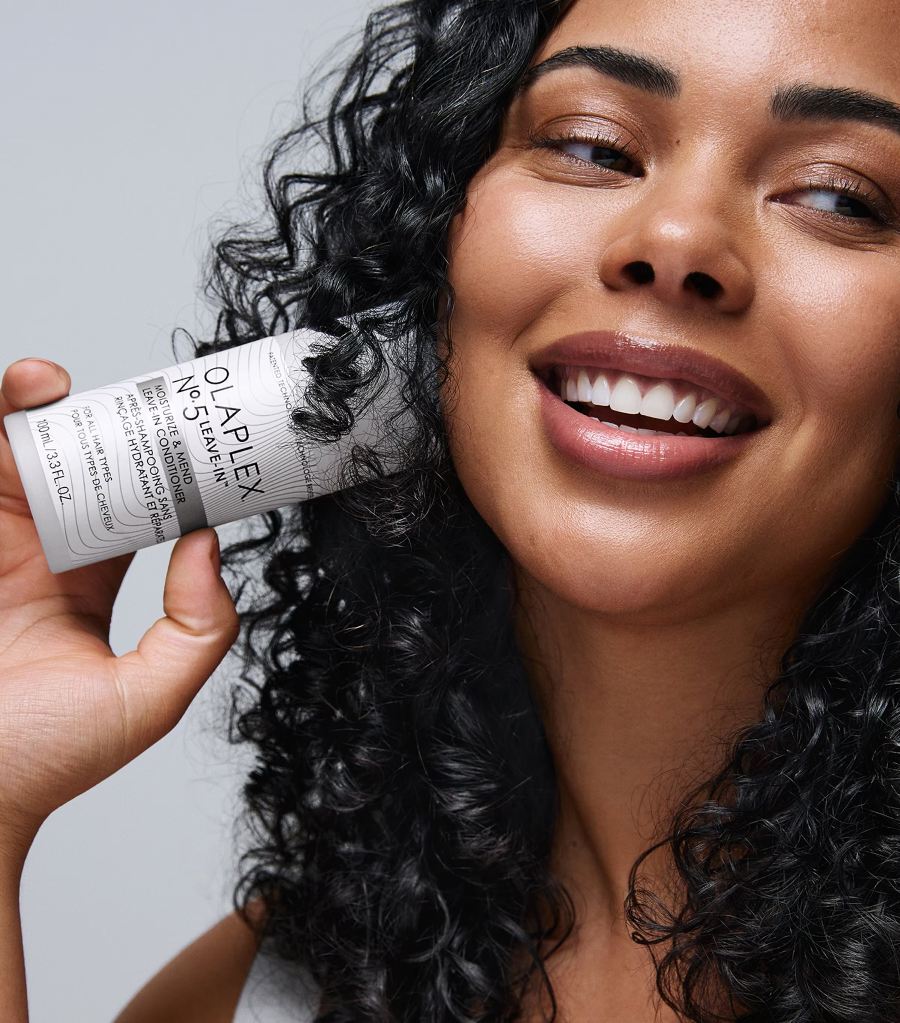 a close up of a woman holding a bottle of olaplex no5. she has curly black hair