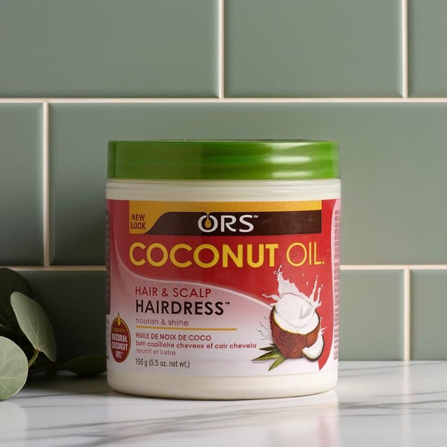 Tub of ORS coconut oil in front of green tiles, on a marble worktop with a small bit of foliage on the left hand side