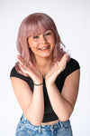A person with ice pink synthetic wavy hair wig posing with hands open and a smile on their face.