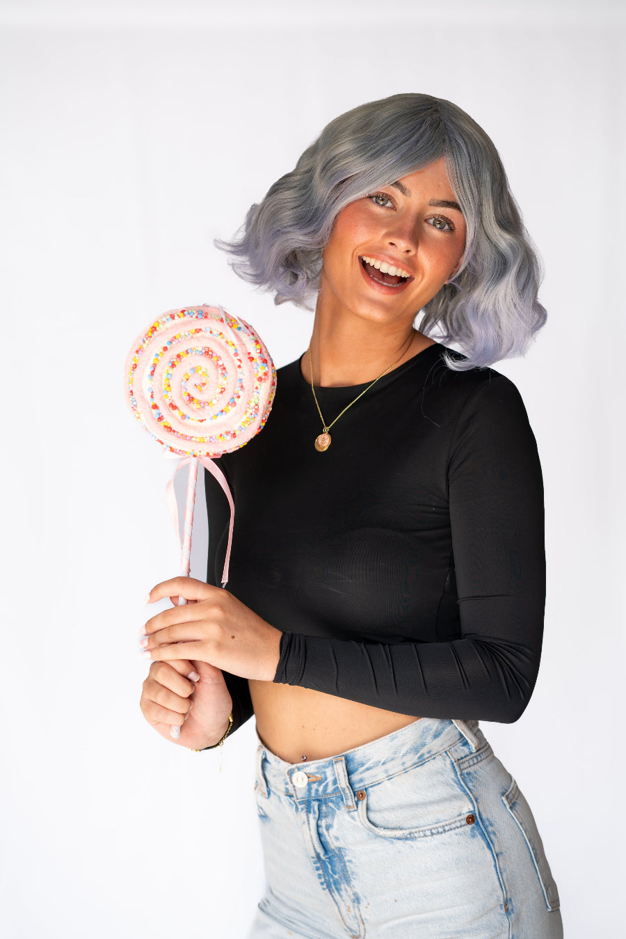 A woman is wearing a synthetic wig with soft curls and grey to light purple color transition, resembling a bob style wig. She is also wearing a black top and blue jeans.