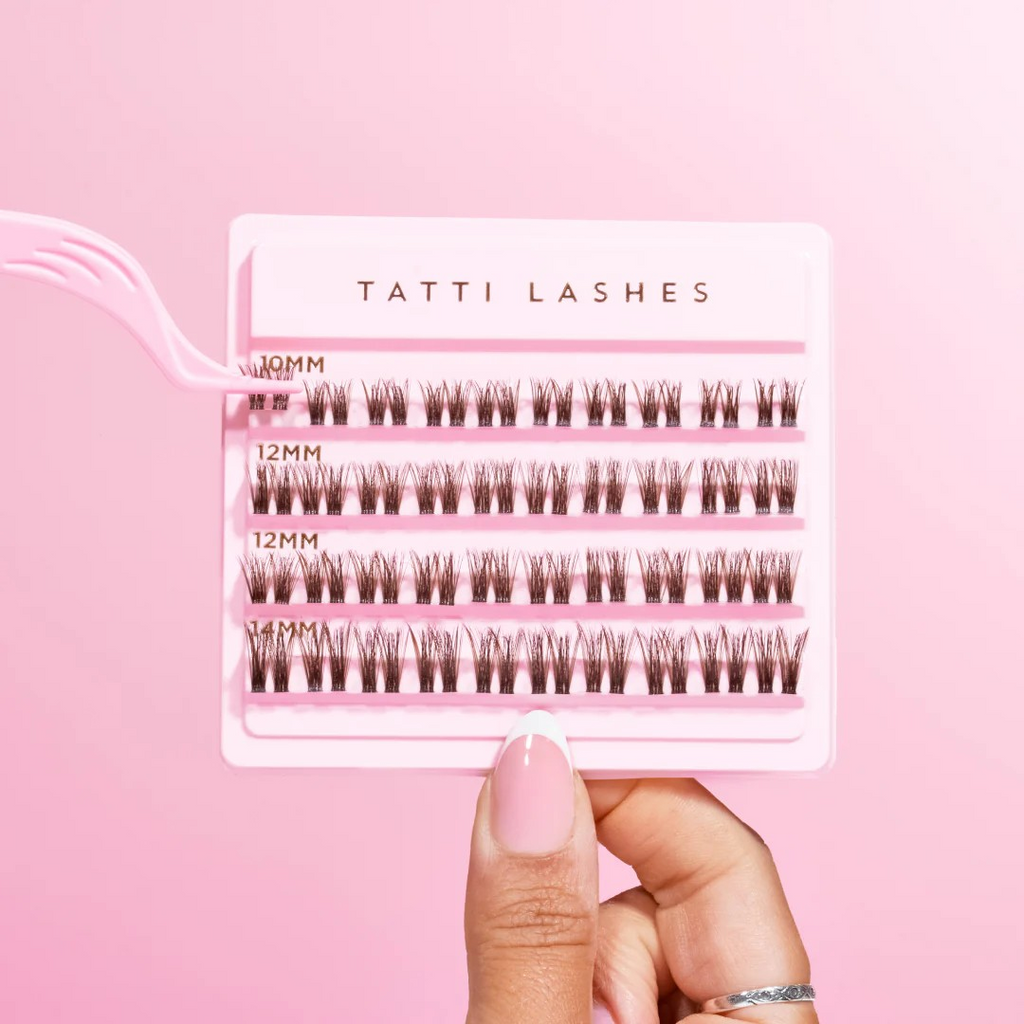 pink box of tatti lash clusters being held up by thumb and finger and a pair of pink tweezers clutching 2 clusters