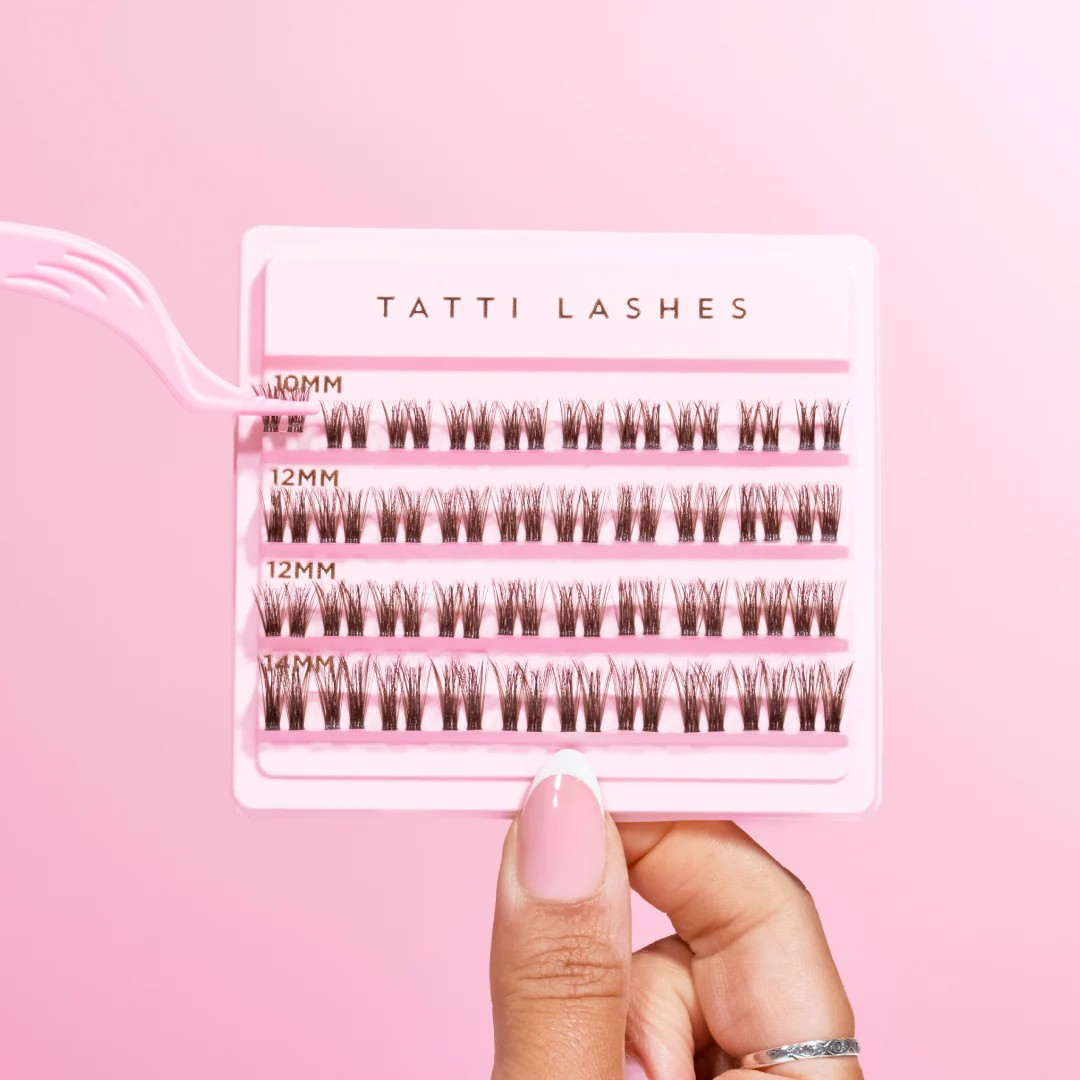 pink box of tatti lash clusters being held up by thumb and finger and a pair of pink tweezers clutching 2 clusters